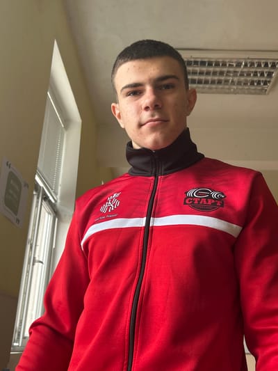 A young male, appearing to be a teenager or young adult, is depicted indoors, looking directly at the camera. He has short, dark hair and a neutral facial expression. He is wearing a red full-zip tracksuit jacket with a black collar and a white horizontal stripe across the chest. On the left side of the jacket, a logo featuring a stylized crown or 'W' symbol is present, with the text "Carlos Nasar" beneath it. On the right side of the jacket, a black circular emblem displays a stylized barbell and Cyrillic text reading "СЪБ", "СТАРТ", and "ЧЕРВЕН БРЯГ". The image is captured from a low angle. The background reveals light-colored walls, with a window fitted with horizontal blinds visible on the left side, admitting natural light. Adjacent to the window on the wall, a paper sign featuring a quote bubble graphic and partially visible Cyrillic text is affixed. The ceiling is light-colored, and a rectangular fluorescent light fixture, appearing incomplete or damaged, is present in the upper right portion of the frame. The location is identified as Cherven Bryag, Bulgaria, based on the text on the individual's jacket.