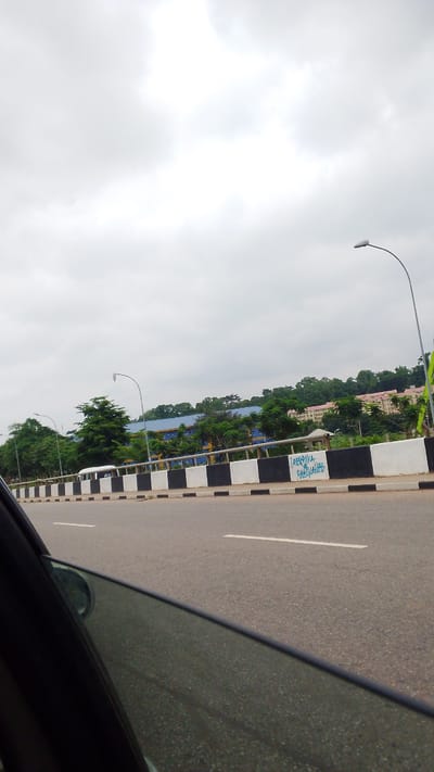 An overcast day in Aso, Nigeria, shows a highway with a black and white striped barrier. Behind the barrier, a white bus is visible amongst trees and buildings. A street light is visible on the right side of the image. The foreground shows the interior of a car with the window partially down.