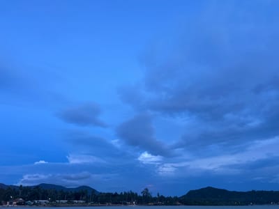 This media file captures a twilight scene over a tropical coastline. The sky is a deep, moody blue, filled with dramatic, textured clouds that suggest an impending storm or the lingering colors of sunset. The lower portion of the image reveals a calm body of water, likely the sea, reflecting the darkening sky.
Along the distant shore, a strip of land is densely populated with palm trees, their silhouettes stark against the sky. Nestled amongst the trees are buildings, possibly homes or resorts, with a few lights twinkling, indicating human presence. The terrain behind the treeline rises into rolling, dark hills or mountains, covered in lush vegetation. The overall mood is serene yet dramatic, evoking a sense of tranquility as day transitions to night, possibly after a day of rain or before a storm. The time of day is clearly evening, given the deep blues and the presence of artificial lights. The location is consistent with a tropical island environment, specifically Ko Pha-ngan District, Thailand, known for its natural beauty and coastal landscapes. No text is visible in the image.