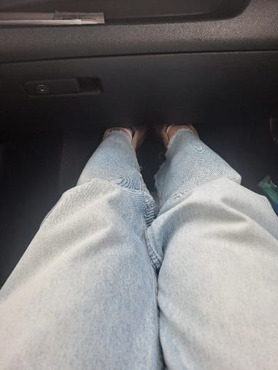 The image displays a first-person perspective captured from within a vehicle interior, looking downward at the occupant's lower body and forward into the dashboard area. The occupant's legs are clothed in light-wash blue denim jeans, which feature frayed or distressed hems above the ankles. The feet are encased in light-colored, open-toe shoes or sandals. Directly ahead, the vehicle's dark-colored dashboard or lower glove compartment panel is visible, characterized by a textured surface. A dark glove compartment latch, incorporating a silver-toned keyhole or USB port, is centrally located on this panel. Above the glove compartment, a narrow horizontal slot contains a white, rectangular item. The floor area beneath the feet appears dark. A partially obscured green or blue object is visible on the far right side of the frame. The specific location in the city of Obukhovo, Russia, is not ascertainable from the visual evidence within the image.