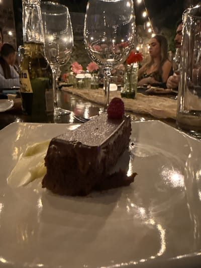 A dark-colored cake with a raspberry on top sits on a white plate at a table. A dollop of cream is near the cake. Wine glasses and a bottle of wine are also on the table. A woman is in the background near floral decorations and lights. The location is likely a restaurant in Santiago de Querétaro, Mexico.