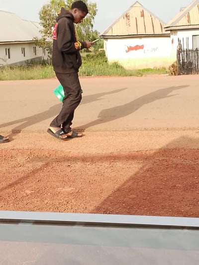 The image depicts a young Black male, appearing to be in his late teens or early twenties, standing on a dirt and asphalt road in Agwala, Nigeria. He is dressed in a black hooded sweatshirt with yellow and red accents on the sleeves and hood, black pants, and black slides. He is holding a light-colored object, possibly a phone or a small notebook, in his right hand, looking down at it. In his left arm, he is carrying a bright green folder.

Behind the individual, there are two buildings. The building on the left is a single-story structure with a white facade and light-colored corrugated metal roofing. It has several windows with visible vertical blinds or louvers. The building on the right also has a white facade and a corrugated metal roof, this one with visible patches and rust. Red paint markings are visible on the wall of this building, with the word "LODGE" clearly legible. A dark, vertical slatted fence separates the property from the road in front of the building.

The environment is an outdoor setting, with dry grass and some trees visible behind the buildings. The road surface is a mixture of reddish-brown dirt and asphalt. Shadows of the person and possibly other unseen objects are cast on the road. The lighting suggests it is daytime, with a bright sky, though it is not possible to determine the time of day precisely. The foreground shows a portion of a gray structure, possibly a low wall or ledge.