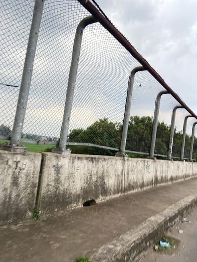 The image shows a chain-link fence along the edge of a concrete bridge in Kecamatan Margaasih, Indonesia. The fence is made of metal and has a rusty brown metal railing along the top. The fence is attached to concrete posts which are secured to the bridge wall. The bridge is made of concrete and has a sidewalk along one side. There is a small drainage hole in the bridge wall. There are green trees and bushes in the background, and a cloudy sky above. The bridge appears to be over a road or highway. The sidewalk is dirty and has some trash on it. The image is taken from a low angle, looking up at the fence.