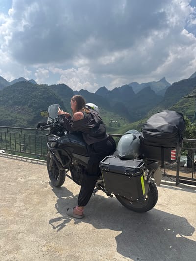 A traveler in dark clothing and sandals is leaning over a motorcycle parked near a metal railing. The motorcycle has luggage, including a large black bag, a gray helmet, and a black side case with a reflective stripe. Behind the motorcycle and rider, there is a mountainous landscape covered in vegetation under a cloudy sky. The photo was taken in Lũng Phìn Commune, Vietnam. The surface beneath the motorcycle appears to be concrete.
