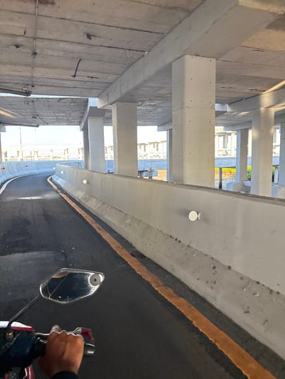 The image depicts a person riding a motorcycle on an elevated road or bridge under construction in South Denpasar, Indonesia. The road surface is asphalt with a visible yellow line marking. The rider's hand and a side-view mirror of the motorcycle are in the foreground. The environment is characterized by unfinished concrete structures, including large pillars and an overhead ceiling. In the background, glimpses of buildings under construction and some vegetation are visible. The lighting suggests it is daytime, possibly late afternoon due to the shadows.