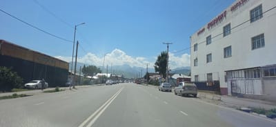 The image shows a street scene in Karakol City, Kyrgyzstan, on a sunny day with a clear blue sky and scattered white clouds. The camera is positioned on a road, likely from a vehicle. On the right side of the road, there is a large, white, multi-story building with a sign that reads "КОМФОРТ" (Komfort) in red lettering. The building has multiple windows on each floor. In front of the building, there is a fence and some enclosed areas. On the left side of the road, there is a long, dark-colored building with a yellow or tan roof.
The road is paved and has double white lines marking the lanes. Several cars are driving on the road, and some are parked along the sides. There are streetlights and utility poles with wires visible along the street. In the background, mountains can be seen, suggesting a mountainous region. There are some trees and vegetation along the sides of the road and in front of buildings.
On the left side of the road, a white car is parked near the dark building, and a person is standing next to it. Further down the street, there are more cars and buildings, creating a typical urban or suburban streetscape. The overall impression is a view of a main road in a town or city, with commercial buildings and residential structures lining the street, set against a backdrop of mountains.