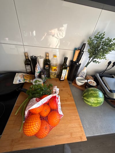 The image depicts a kitchen countertop scene within a residence in Remscheid, Germany. A large rectangular wooden cutting board occupies the central foreground, featuring a bunch of fresh green cilantro and a red mesh bag holding several oranges. To the left of the cutting board, a black frying pan rests on a stove burner, and a package of "Lidl Pimientos Padrón" (frozen Padrón peppers) is partially visible, adjacent to a white-labeled can.
Along the rear of the countertop, a collection of kitchen items is arranged. From left to right, these include three dark glass bottles of olive oil, one clear plastic bottle of sunflower oil, a black pepper grinder, a white salt shaker, and a black cylindrical knife block containing several knives. To the right, a small white vase with blue patterns holds a sprig of artificial green foliage. Adjacent to this, on a smaller wooden board, are a white ceramic mortar with a pour spout, a small jar labeled "Himalaya Salz" (Himalayan salt), and a small, striped watermelon. An electric grill/sandwich maker, silver and black, is positioned on the far right, connected to a white wall outlet.
The background consists of reflective, white, high-gloss wall tiles or cabinet fronts, which clearly display the reflection of a person, likely a child, wearing a light-colored long-sleeved top. A foot wearing a light grey sock is visible at the bottom center of the frame, indicating the presence of an individual standing at the counter.