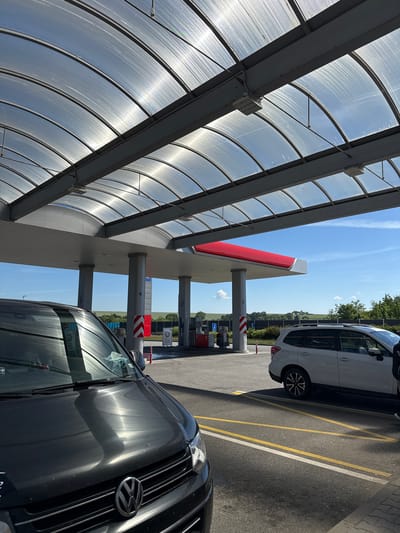 The image depicts a gas station canopy under a clear blue sky with scattered clouds. The canopy is constructed with a translucent, corrugated plastic roof supported by metal beams, creating a series of arched segments. Beneath the canopy, a row of gas pumps is visible, with some displaying fuel prices. Two cars are present: a dark-colored van in the foreground, and a white SUV to the right, positioned at a gas pump. The ground is asphalt, marked with yellow and white lines indicating parking or driving lanes. In the background, a grassy field with distant trees and rolling hills suggests a rural or suburban setting outside the city. The scene is brightly lit by sunlight, casting reflections on the cars and the canopy. There are no visible people interacting with the vehicles or the gas pumps. The location is identified as a gas station in Mitrovice, Czechia.