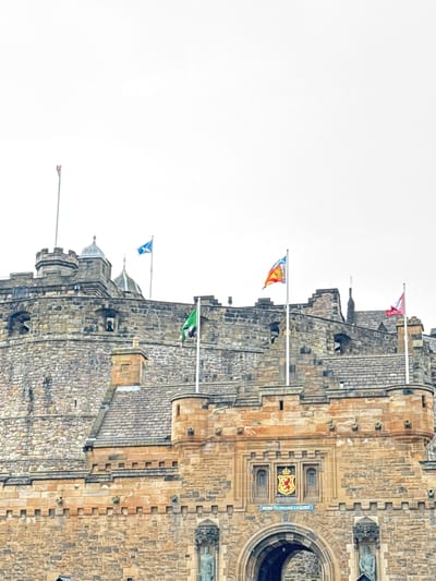 This image shows a low-angle, full-frame view of Edinburgh Castle, located in the city of Edinburgh, United Kingdom. The castle is constructed primarily of stone, with visible turrets, battlements, and architectural details. Several flags are flying from poles on the castle walls. One flag, on the left, is blue with a white diagonal cross, representing the Scottish flag. To its right, a multi-colored flag with red, yellow, and blue elements is visible. Further to the right, a red and white flag is flying. A green flag with a crest is also prominent on the left side of the image. Below the flags and in the central portion of the image, a large stone archway is visible, leading into the castle. Above the archway, a shield with a red lion rampant on a gold background is displayed. A Latin inscription, "NEMO ME IMPUNE LACESSIT," is visible below the shield. The sky is overcast and white, suggesting a cloudy day. The overall scene depicts a historical landmark under a subdued sky.