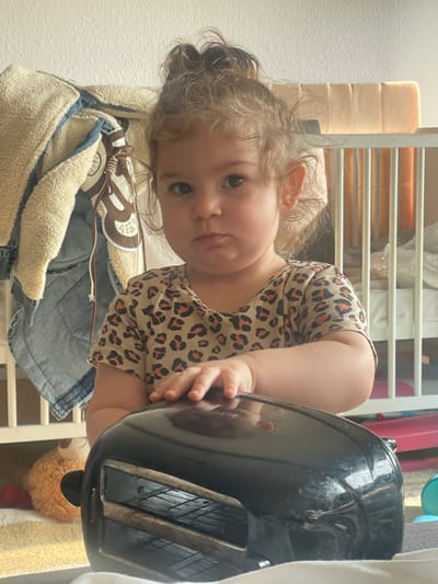 The image captures an indoor scene in Senden, Germany, featuring a toddler-aged child positioned in front of a black toaster oven. The child has light brown, curly hair pulled into a top knot, wears a short-sleeved, leopard-print shirt, and has a small gold earring in the visible right ear. Their expression is a slight pout as they look directly at the camera. The child's right hand rests on the reflective, black top surface of the toaster oven, which displays interior wire racks through its front opening. In the background to the left, a beige-lined denim jacket hangs, partially obscuring a stuffed animal and red toys on a striped rug. To the right, a white wooden crib with light-colored padding is visible. The foreground indicates a soft, light-colored surface, possibly a blanket or rug.