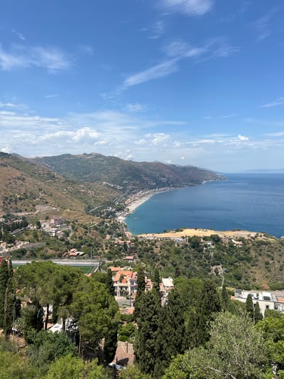 An aerial, high-angle, eye-level, long shot captures a sunlit, panoramic view of the coastal city of Taormina, Italy. The blue sea, dotted with white-capped waves, meets a vibrant green, hilly coastline. A long, sandy beach stretches along the shore, with a vibrant blue, turquoise sea on its right. Buildings, painted in earthy tones of brown, orange, and white, are scattered across the landscape, nestled amongst lush green trees and olive groves. A winding road snakes through the hills, connecting the various parts of the city. In the distance, mountains rise majestically, their peaks disappearing into a hazy sky. The sky itself is a brilliant blue, punctuated by scattered white clouds. The scene is peaceful and tranquil, with no people or animals visible. The overall impression is one of natural beauty and serene coastal living.