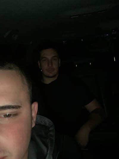 In a dimly lit indoor environment, two individuals are present. The person in the foreground is partially visible, with their left eye and cheek the most prominent features, their gaze directed towards the camera. They are wearing a dark gray hooded garment. Behind them, a second individual is more fully depicted, facing forward with a neutral expression. This person is wearing a black t-shirt. The background is dark and indistinct, suggesting it may be the interior of a vehicle or a room with minimal lighting. There are no discernible landmarks or contextual clues to indicate a specific location within Yasen, Bulgaria.
