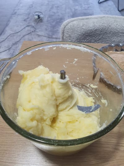 A close-up shot shows mashed potatoes inside a glass food processor bowl. The potatoes are a pale yellow and appear smooth, with some slightly lumpy textures. A metal blade is visible within the bowl, embedded in the mashed potatoes. The bowl rests on a light brown wooden surface. In the background, a blurred gray surface with black cords is visible, possibly a floor with electrical outlets or cables. A lightly textured gray object, possibly a piece of furniture or fabric, is also present. The lighting is warm and somewhat diffused. The scene is indoors. This image is not related to Kheyrabad, Iran.