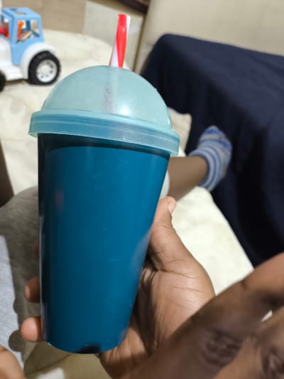 A dark-skinned hand grasps a teal-colored, opaque plastic tumbler. The tumbler is topped with a clear, light blue dome lid, through which a red and white striped plastic straw protrudes vertically. The tumbler appears to contain a dark liquid. To the upper left, a portion of a white and light blue plastic toy truck is visible on a light-colored surface. To the right, a foot wearing a light blue and white striped sock rests on a light-colored upholstered surface. A dark blue fabric is partially visible behind the foot. The scene is indoors, located in Nicosia, Northern Cyprus / Cyprus.