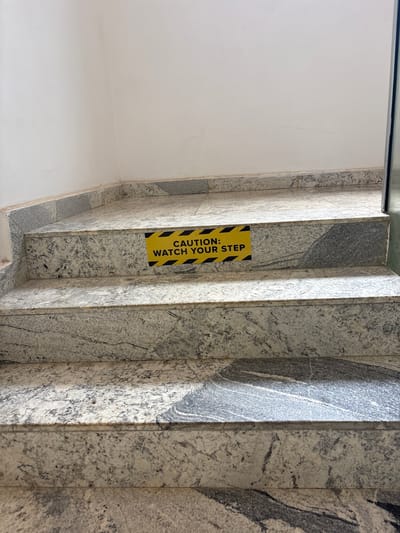 The image shows a close-up view of a granite staircase in Gwaska, Nigeria. A yellow caution sign with black hazard stripes is affixed to the second step from the top, reading "CAUTION: WATCH YOUR STEP". The stairs are made of speckled grey and white granite, with some darker veining. The wall above the stairs is painted white. The scene appears to be an indoor staircase in a building. There are no people or animals visible in the image. The lighting suggests it is daytime.