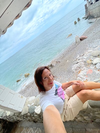 A woman with brown hair and a white t-shirt with a colorful graphic print is taking a selfie on a rocky beach. She is wearing light-colored shorts and is seated with her legs crossed. Behind her, the turquoise ocean stretches to the horizon under a cloudy sky. Concrete barriers are visible in the water and on the shore, suggesting coastal defenses or remnants of a structure. The beach is covered in small, grey pebbles and larger rocks. Part of a white, brick-like structure is in the foreground, and a textured stone wall is below it. The scene appears to be in Abaata, Abkhazia.