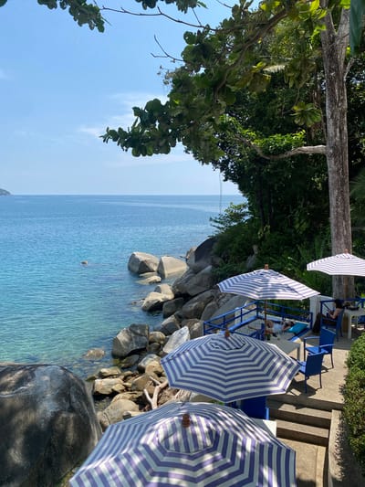The image captures a coastal scene at Kamala, Thailand. The view is from an elevated position looking toward the sea. In the foreground, there are partially visible beach umbrellas with blue and white stripes. Below the umbrellas, rocks and pebbles form the coastline. The sea extends into the distance, with varying shades of blue. A small, light-colored object is visible in the water. On the right, there are stairs and blue chairs. On the left, tree branches and foliage are visible, contrasting with the blue sky. People are visible beneath one of the umbrellas, some seated.