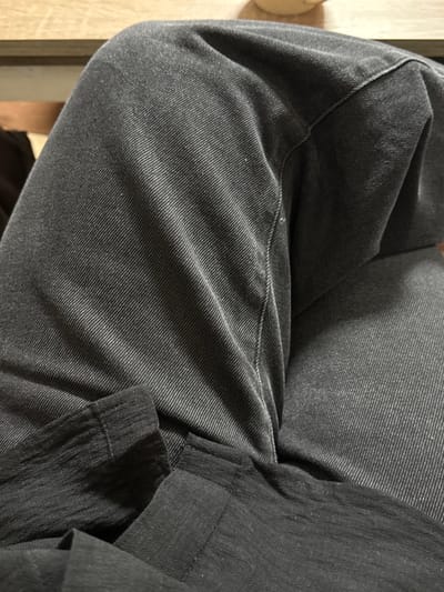 A close-up shows the texture of a dark gray garment, likely pants. The fabric appears to be a ribbed corduroy, creased and folded, suggesting wear. To the right, a contrasting, darker fabric, possibly linen, drapes over the corduroy, adding depth and visual interest. The lighting accentuates the textures, highlighting the fine details of each material.