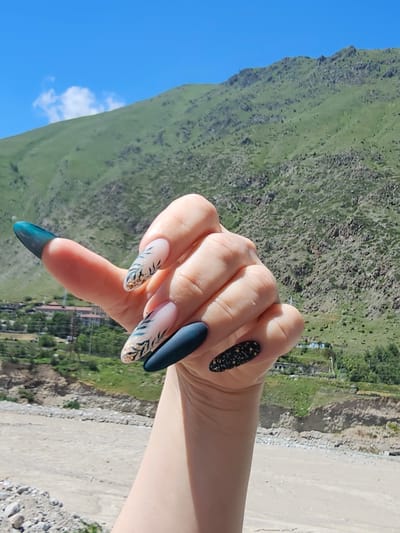 A hand with elaborately decorated fingernails is held up in front of a mountainous landscape under a clear blue sky with a few wispy clouds. The hand features long, almond-shaped nails, with designs including a gradient of teal and dark blue on the thumb, a matte dark blue on one finger, and two nails adorned with a light pink base, dark green leaf patterns, and gold flecks. The background shows a lush green mountainside, a dry riverbed, and a small settlement in the distance, likely in the city of Tyrnyauz, Russia, given the mountainous terrain and settlement structure. The lighting suggests it is daytime, with the sun casting subtle shadows.