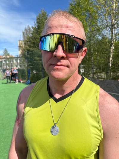 A man with light skin, short blonde hair, and wearing mirrored sunglasses, is pictured in a close-up, low-angle shot. He is wearing a light green tank top with black trim and a silver dog tag necklace. The background shows trees and a building in the distance. There is a green surface, possibly artificial turf, in the immediate background. Blurred figures of people sitting on chairs are visible in the background. Based on the description of Polarnie Zori, Russia, it's possible the photograph was taken outdoors during daylight hours in a park-like setting.