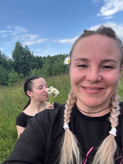The image shows two women in a field with trees in the background and a blue sky with scattered clouds above. The woman in the foreground is taking a selfie and smiling at the camera. She has blonde hair in two braids and a daisy tucked behind her ear. The woman in the background is holding a bouquet of daisies and smelling them with her eyes closed. She has dark hair in a braid. The location appears to be a rural or natural area, possibly a meadow or field on the outskirts of Pozdyshki, Russia.
