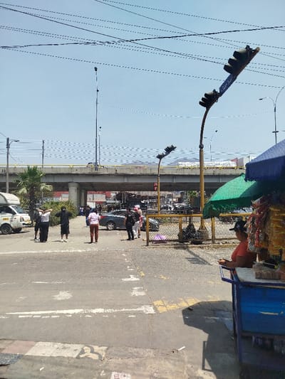 A street scene captures several individuals gathered near a crosswalk, with a bridge and a traffic light fixture visible in the background. A woman is seen sitting behind a vendor's cart. Power lines stretch across the blue sky, and the bridge has a yellow railing. Cars are parked nearby, and there appears to be a security or police presence within the group of people.