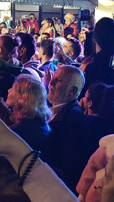 The image depicts a dense gathering of people at an outdoor or semi-outdoor event, likely taking place at night or in the evening in Ash-Shaghour Municipality, Syria. The scene is illuminated by artificial lights, casting a mix of purple, blue, and warm tones across the crowd.

In the foreground, an older man with a white beard and receding hairline is prominently featured in profile, looking upwards and to the right, appearing deeply focused. He is wearing a dark jacket over a white collared shirt. To his left, a woman with light-colored, curly hair is also seen in profile, facing left. An arm with a black beaded bracelet is visible in the lower left corner, indicating the proximity of the viewer to the crowd.

The midground and background are filled with numerous other individuals. Many attendees are holding up smartphones, suggesting they are recording or photographing a central event or performance that is not visible within the frame. Several children are interspersed throughout the crowd, some appearing to be on shoulders or elevated platforms for a better view, including a young girl with braids in a red and white jacket and another child wearing a red jacket with a furry hood.

The background shows decorative elements, possibly scalloped awnings or temporary tent-like structures, implying a festive or market-like atmosphere. The collective attention of the crowd and the widespread use of cameras indicate an engaging public spectacle or entertainment is unfolding. There is no visible text in the image.