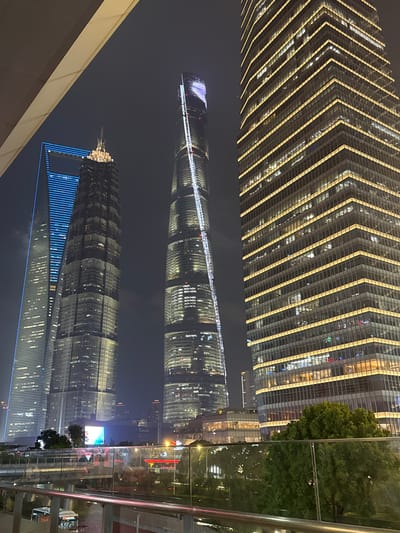 The image depicts a nighttime view of a modern cityscape, likely in Pudong, China, with a focus on towering skyscrapers. The perspective is from an elevated vantage point, possibly a walkway or balcony, looking down and across at the illuminated buildings.
The most prominent subject is a collection of skyscrapers, characterized by their sleek, glass facades and intricate lighting designs. One building in the foreground appears to be a particularly tall, curved structure, reflecting the lights and the night sky. Another skyscraper, partially visible, has a distinctive patterned facade with horizontal yellow lighting. Further in the distance, a building is illuminated with bright blue vertical lines.
The time of day is clearly night, with the darkness of the sky contrasting with the vibrant artificial lights of the city. There are no discernible people visible in the image, and the primary objects are the architectural structures of the skyscrapers. The scene is urban and futuristic, showcasing advanced architecture and urban planning.
Notable details include the reflections of the lights on the glass surfaces of the buildings, creating a dazzling visual effect. The presence of trees suggests a landscaped urban environment, possibly a park or plaza, interspersed among the concrete jungle. The overall mood conveyed is one of grandeur and modernity, highlighting the dynamism of a major metropolitan area at night. No visible text is identifiable in the image.