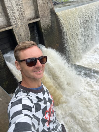 The image shows a man taking a selfie at a waterfall. The man is wearing sunglasses and a black and white checkered shirt. He is smiling and appears to be happy. The waterfall is cascading down behind him, creating a misty spray. The scene is outdoors, and the weather appears to be overcast. The man's clothing and the background suggest a casual outing, possibly a tourist visiting a natural attraction. There is no visible text in the image.