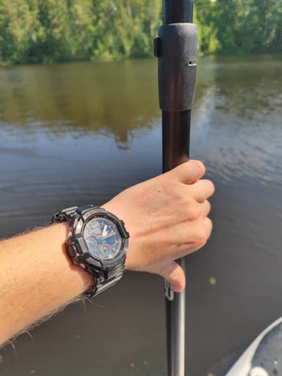 A close-up, high-angle shot shows a person's left arm and hand holding a black paddle on a body of water. The person is wearing a black G-Shock watch on their wrist. The background features a blurred green tree line and the dark, rippling surface of the water.