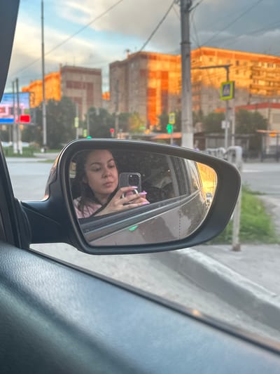 The media file captures a reflection in a car's side mirror, showing a young woman taking a selfie with her phone. She is looking at the camera, with one hand holding the phone and the other, with purple-painted nails, is visible near her face. She has dark hair and is wearing a pink shirt. The reflection also shows the interior of the car, including the window and part of the dashboard.
Outside the car, as reflected in the mirror and visible in the rest of the image, is an urban scene. Tall, multi-story buildings with orange and brick facades dominate the background, suggesting an Eastern European city. A street with a sidewalk and traffic lights is visible. One traffic light displays a green signal. There are also utility poles and wires, common in urban environments. The sky is partly cloudy. The warm, golden light suggests it is either early morning or late afternoon. The overall mood of the selfie is casual and relaxed. The location context provided is Magnitogorsk, Russia.