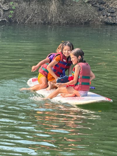 Two young girls wearing life vests stand on a white paddleboard in a body of water. The water reflects light and creates wavy patterns. The background shows vegetation along the shoreline. This appears to be Tambon Nam Phrae, Thailand.