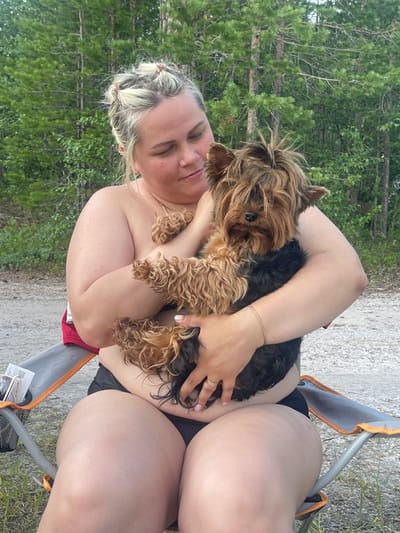 A woman with blonde hair tied up in buns is sitting in a folding chair outdoors and holding a Yorkshire terrier. She is wearing black bikini bottoms and is shirtless. The dog is brown and black with shaggy fur. They are in a wooded area with tall pine trees in the background. The ground is covered in gravel and some grass. There is a folding chair next to the woman with a small bag on it.