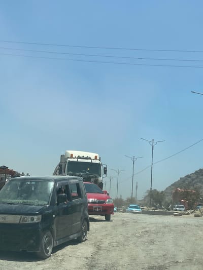 The image depicts a dusty, sunlit outdoor scene, likely on a road within the city of Kokran, Afghanistan. In the foreground, a black Suzuki Wagon R is parked or moving slowly, with its driver visible in the driver's seat. To the right of the black car, a red sedan is also present. Behind these vehicles, a white truck is stopped or moving slowly. The road surface appears to be unpaved and rough. In the background, there are several utility poles with streetlights, some trees, and a rocky, mountainous terrain. The sky is clear and blue, with a few overhead wires visible. The overall impression is one of a busy, possibly traffic-congested, environment in a semi-arid or mountainous region.