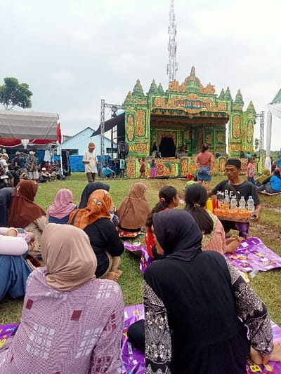 The scene takes place in an open-air area in Kecamatan Margorejo, Indonesia, located at coordinates -6.735 latitude and 111.016 longitude. A large, elaborate stage adorned with ornate carvings and colorful decorations is the focal point, with a sign reading "Happy Birthday." The stage is backed by a tall, thin radio tower. Several people are gathered around the stage, including a group of women and children. Many women are wearing headscarves and traditional clothing. A man is standing near the stage with a tray of drinks. He is facing the camera and appears to be selling refreshments. The ground is covered in grass and a few people are sitting on purple plastic mats. The scene is casual and relaxed, likely a community event or celebration.