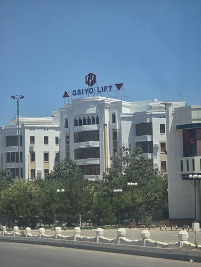 The image depicts a modern building with a clear blue sky in the background. The building is white with several floors and numerous windows. A large sign with blue letters spelling out "OSIYO LIFT" is prominently displayed on the upper part of the building. The sign also features a red triangular logo.
To the right of the building, a sidewalk with decorative concrete bollards separates the building from a paved road. Several lampposts with multiple lights are visible along the sidewalk and in front of the building, suggesting an urban or commercial setting. Lush green trees are interspersed around the building and along the sidewalk, providing a contrast to the architectural elements.
The orientation of the image is rotated, with the top of the building appearing on the left side and the sky on the right. The lighting suggests it is daytime, likely a sunny day given the clear sky. The presence of the "OSIYO LIFT" sign indicates a business or service establishment. The overall scene conveys a sense of development and modern infrastructure in Bukhara, Uzbekistan. No people are visible in the frame.