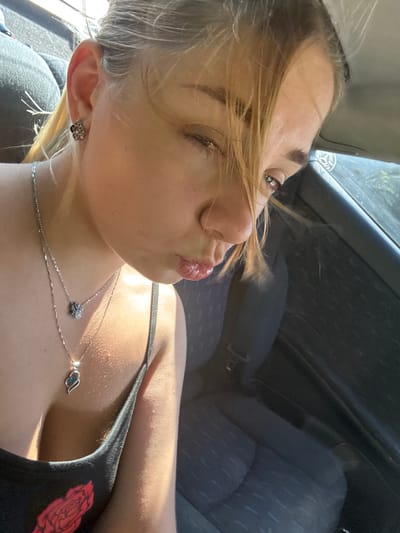 The image shows a close-up, low-angle shot of a young woman with blonde hair sitting in a car. She is looking towards the camera with one eye partially closed and her lips pursed. She is wearing a black tank top with a red rose design and two silver necklaces, one with a small cluster pendant and another with a heart-shaped pendant with a blue stone. She also has a small square earring in her left ear. The interior of the car is visible, including a black car seat. The lighting suggests it is daytime, possibly with sunlight coming from the right side of the image. No specific location in Sredets, Bulgaria is identifiable from the image content.