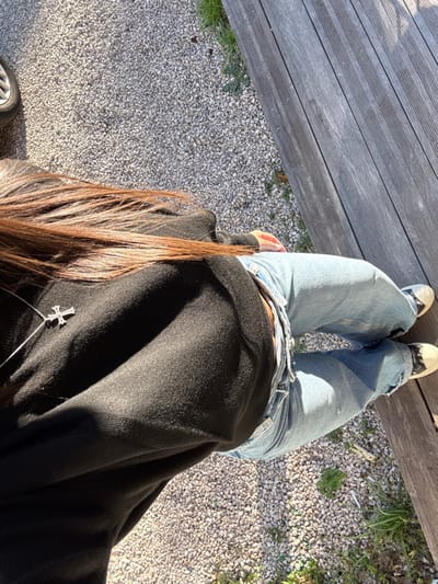 An overhead view depicts a person standing outdoors on a sunny day. The individual, seen from the upper back down to the feet, has long, brown hair cascading down their back. They are wearing a loose-fitting black long-sleeved top and light blue, distressed denim jeans. A silver-toned chain with a small cross pendant is visible against the black top. The person's footwear consists of light-colored low-top sneakers with dark soles or accents.

The immediate ground surface is composed of light grey gravel or small stones, with sporadic patches of green grass and weeds visible, particularly along the edges. To the right of the person, a weathered wooden deck or platform made of parallel planks is present. In the upper left corner, a portion of a car tire with a silver rim is visible. A discarded cigarette butt lies on the gravel near the wooden deck. Direct sunlight creates distinct shadows across the scene. The image contains no discernible visual markers or landmarks to confirm the location as Antibes, France.
