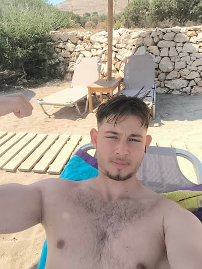 A young man with short, dark, messy hair and a light beard takes a selfie on a beach in Chalki, Greece. He is shirtless and flexing his bicep, with a blue towel draped over the lounge chair he is sitting in. In the background, there are more lounge chairs, a small wooden table, a stone wall, and vegetation. The beach is sandy.