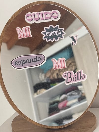 The image shows a round mirror with a wooden frame, reflecting a partially visible scene. The mirror is adorned with stickers in Spanish, reading "Cuido Mi energia," "A Mi Brillo," and "expandiendo." The reflection includes a closet with clothing items and a partially visible interior of a room. The presence of Spanish text on the stickers suggests the location is likely in a Spanish-speaking country. The city of Medellín, Colombia, is not evident from this image alone.
