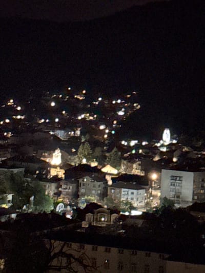 The image depicts a nighttime scene in Sveti Konstantin, Bulgaria. A street with lit streetlights is visible, lined with buildings and trees. The buildings are a mix of residential and commercial structures. Some buildings have lit windows. The overall ambiance suggests a calm, urban neighborhood setting.