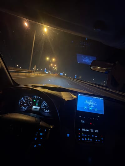 The image shows the interior of a car at night, taken from the driver's perspective, while driving on a well-lit road or bridge in Umm Salal Muhammad, Qatar. The scene is dominated by the dark interior contrasted with the bright dashboard displays and exterior streetlights.
Inside the car, the illuminated dashboard shows an odometer reading of "88888" and a trip meter of "586". To the right, a large infotainment screen displays that the song "Tantra" by "Miyagi & Andy Panda" is playing, with the current time being "7:36". The climate control settings on the screen indicate an "OUTSIDE TEMP" of "35" degrees Celsius, while the dual-zone interior temperature is set to "22.0" degrees Celsius. Various buttons are visible on the console, including "ENGINE START STOP", "HOME", "EJECT", "VOL", "AUTO", "OFF", and controls for "FRONT", "REAR", and "A/C". The steering wheel also features illuminated control buttons, including "ENTER" and directional arrows.
Through the windshield, a multi-lane highway or bridge is visible, brightly illuminated by a series of yellow streetlights extending into the distance. The road appears clear, suggesting smooth night driving conditions. The rearview mirror shows a blurred reflection of the exterior lights.