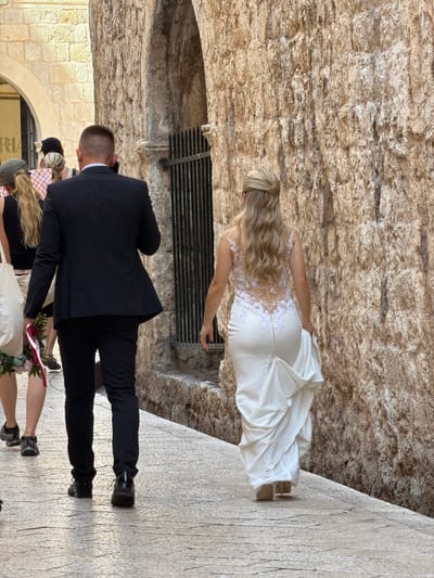 In the historic city of Dubrovnik, Croatia, a newlywed couple is captured walking away from the camera on a stone-paved pathway. The groom, dressed in a black suit and shoes, walks ahead, while the bride, in a white wedding dress with an intricate lace back and a train trailing behind her, follows closely. Other individuals, partially visible, are also on the pathway. The scene unfolds against a backdrop of ancient stone walls and an arched gateway, characteristic of Dubrovnik's Old Town.