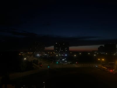 The image depicts a night scene in Domodedovo, Russia, with an urban landscape characterized by buildings and roadways illuminated by artificial lights. Two tall apartment buildings with illuminated windows are prominent in the mid-ground. A multi-lane road runs through the scene, with streetlights casting pools of light. Traffic signals are visible, with some displaying green lights, indicating traffic flow. Several vehicles, including cars and what appear to be trucks, are parked or moving along the road and in parking areas. The sky is dark, suggesting it is late evening or early morning, with some faint clouds visible near the horizon, tinged with the last light of sunset or the first hint of sunrise. The overall impression is one of a typical urban environment at night, with the infrastructure and activity associated with a residential and commercial area.
