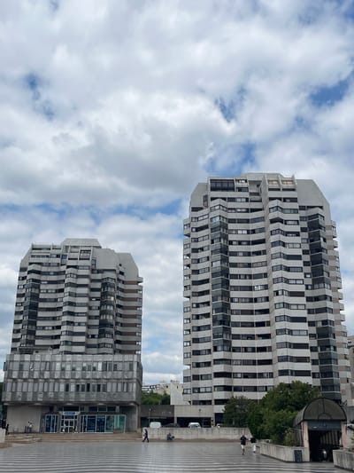This image shows a view of two modern, angular apartment buildings in Créteil, France. The buildings are tall and made of concrete and glass, with a repeating pattern of windows that give them a textured appearance. The building on the right is closer to the foreground and appears to be part of a complex with a lower section that has an entrance and a curved roof structure. A few people are visible walking on the paved area in front of the buildings, some are also visible in the background. One person is walking to the right on the pavement, and another is sitting on a ledge. A white van is parked near the entrance of the building on the right. The sky is filled with scattered clouds, suggesting it is daytime, possibly late morning or afternoon. The lighting is diffused, indicating it might be partly cloudy. The perspective is from a low angle looking up at the buildings, emphasizing their height and imposing architecture. There are no discernible emotions conveyed in the image, which focuses on the architectural elements and the urban setting.