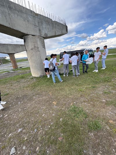 A group of eight males stand next to a white van under a partially constructed concrete bridge structure with rebar protruding from the top. The van is parked on a grassy area. The men are all wearing casual clothing; most are wearing white shirts. One male is carrying a white box and another is wearing a light blue shirt. The background features a vast field with a cloudy sky above. The location is Getk, Armenia.