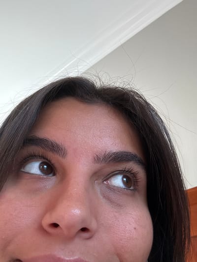 The image displays a close-up, upward-angled view of a person's upper face, specifically a female with dark brown hair and warm-toned skin. The subject's dark brown hair frames the forehead, with visible flyaway strands at the crown. Both dark brown eyes are directed upwards and slightly to the viewer's right, revealing a portion of the sclera on the inner sides. Long, dark eyelashes are prominent, and the eyebrows are thick and well-defined. The bridge and tip of the nose are visible below the eyes, along with the upper lip and a hint of the lower lip at the very bottom edge of the frame, suggesting a subtle upward curve. The skin on the forehead and cheeks exhibits texture. The background consists primarily of a plain white or off-white ceiling, featuring a section of decorative crown molding in the upper left corner. A dark brown, vertical object, possibly a wooden door or piece of furniture, is visible along the far right edge of the frame. The scene is situated indoors, within an unspecified room located in Princeton, United States.