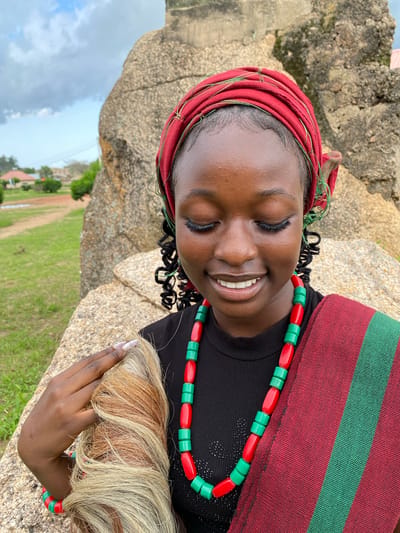The media file shows a young Black woman with her eyes closed and a smile on her face, leaning against a large rock. She is wearing a red headwrap adorned with thin green threads, a black top, and a beaded necklace made of alternating red and green beads. A red and green striped cloth is draped over her shoulder. Her right hand is holding a bundle of light-colored hair. The scene appears to be outdoors, with a grassy field and some trees visible in the background. The sky is cloudy. The image is oriented vertically. This appears to be a portrait of the woman in a natural setting, possibly in Wang, Nigeria. The lighting suggests it is daytime.