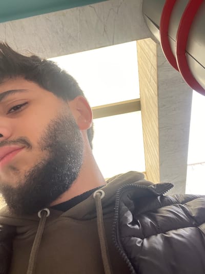 A man in a dark puffer jacket and green hoodie faces the camera. The perspective is unusual, shot from below and slightly to the side, causing the scene to appear rotated. He has dark hair and a full beard. The background includes a grey marble wall, a window letting in bright, diffused sunlight, and architectural details with red accents on a grey surface. The lighting is strong and somewhat obscures details.