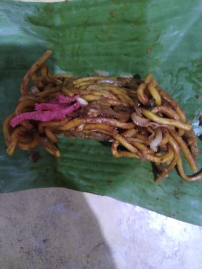 A serving of stir-fried noodles is presented on a large, deep green banana leaf in Gampongbaro, Indonesia. The noodles are thick, yellow-orange, and coated in a dark reddish-brown sauce, appearing glossy. Interspersed among the noodles are translucent white slivers, consistent with sliced onions, and a distinct cluster of bright pink, shredded or stringy material, likely a traditional topping or condiment. The banana leaf itself exhibits minor creases and some residue, suggesting it has been used to hold the food. The leaf rests on a light grey, textured surface, possibly concrete or a stone slab. No individuals are visible in the scene.